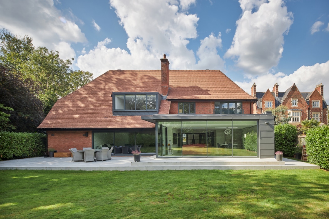 A modern brick house with a red tile roof and large glass extension, featuring a patio with outdoor seating and a lawn, surrounded by trees and neighboring houses.