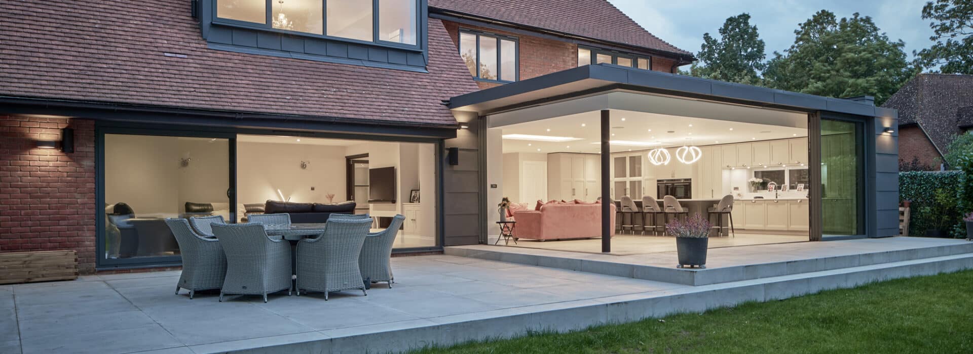 Modern house with large glass sliding doors opening onto a patio with outdoor dining furniture and well-lit interior kitchen and living areas.