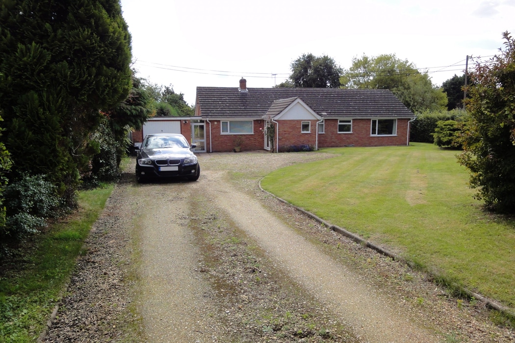 Single-story brick house with a gabled roof, surrounded by a large lawn and trees. A black car is parked on a gravel driveway leading to the house and attached garage.