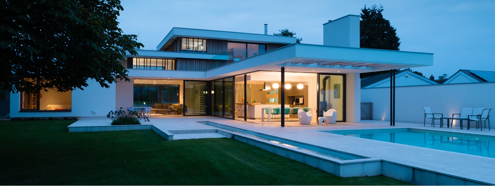 Modern two-story house with large glass windows, outdoor patio, pool, and lawn, illuminated at dusk.