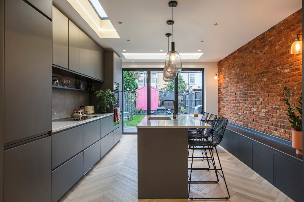 Modern kitchen with grey cabinets, an island with bar stools, exposed brick wall, pendant lights, and large glass doors opening to a garden with a pink shed.