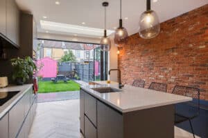 Modern kitchen with a marble island, pendant lights, exposed brick wall, and sliding glass doors opening to a backyard with a pink shed and picnic table.