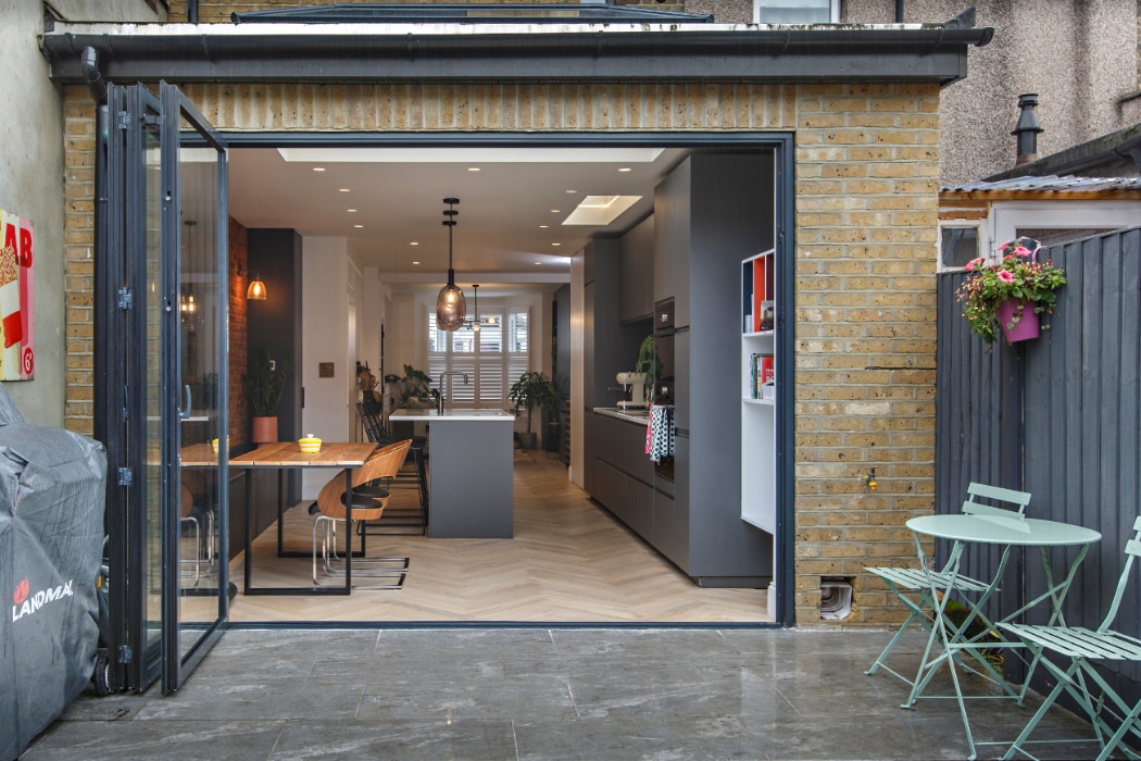 Modern kitchen and dining area with open glass doors to a patio featuring a small table and chairs; brick exterior and herringbone wood floor visible.