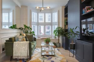 A modern living room with a green sofa, glass coffee table, patterned rug, indoor plants, large bay window with white shutters, and built-in dark shelving unit.
