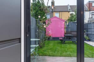 View through a glass door showing a garden with a green lawn, a pink shed, a black picnic table, and a gray fence.