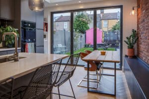 Modern kitchen with island, wire bar stools, wooden dining table, and sliding glass doors opening to a backyard with a pink shed and outdoor furniture.