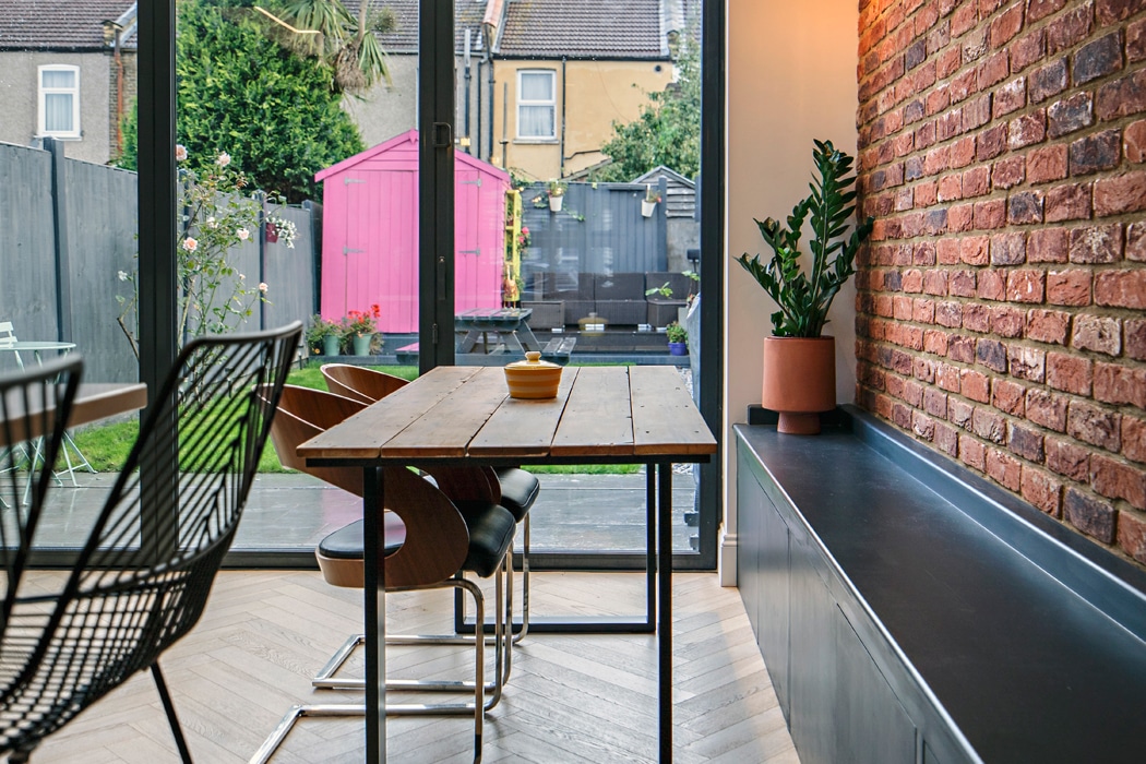 Modern dining area with a wooden table and chairs next to a brick wall, overlooking a backyard with a bright pink shed through large glass doors.