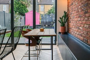 A modern dining area with a wooden table and metal chairs next to glass doors overlooking a backyard with a bright pink shed and plants.