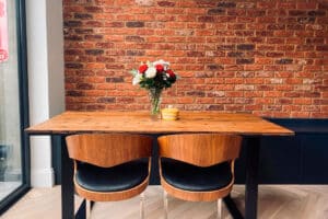 A wooden table with two chairs sits against a brick wall, topped with a vase of flowers and a small yellow bowl.