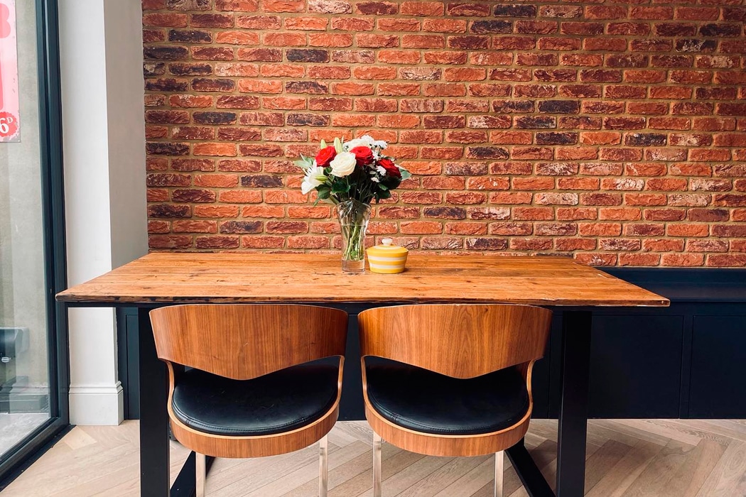 A wooden table with two chairs sits against a brick wall, topped with a vase of flowers and a small yellow bowl.