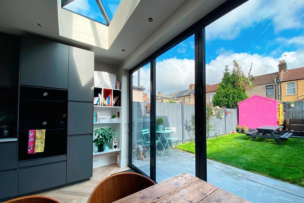 Modern kitchen with black cabinets, wooden dining table, and large glass doors opening to a backyard with green lawn, pink shed, and outdoor seating under a blue sky.
