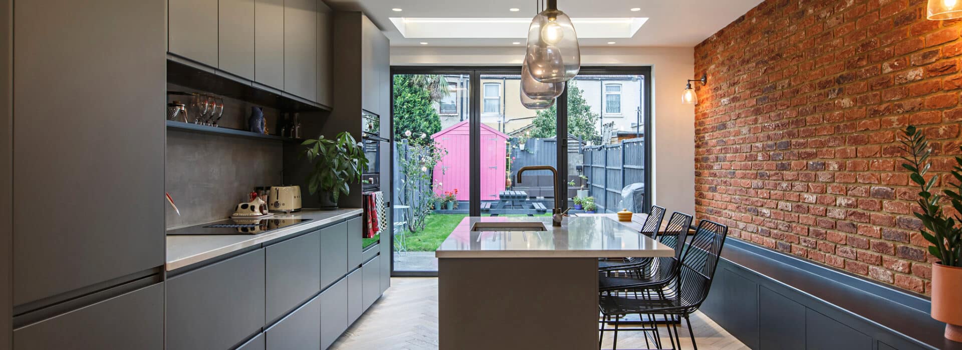 Modern kitchen with premium glazing bifold doors opening onto a garden terrace, featuring sleek cabinetry and exposed brick walls.