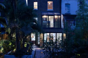 A modern, two-story brick house with large glass windows and doors illuminated at night, surrounded by a lush, landscaped garden with tall plants and trees.