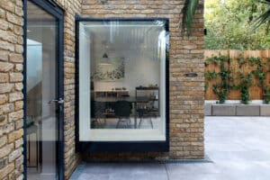 Large glass window protruding from a brick house wall, revealing a modern dining area inside; patio with gray tiles and wooden fence outside.