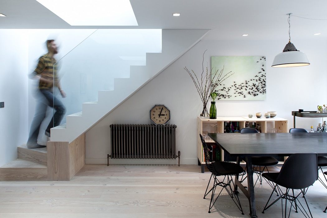 A person ascends a modern staircase in a bright dining area featuring a dark table, black chairs, wall art, a clock, and minimalist decor.
