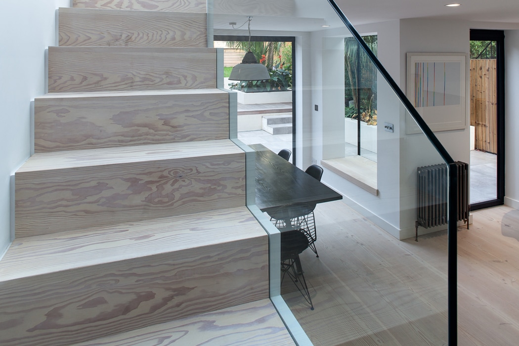 Modern interior with wide light wood stairs and glass railing leading to an open dining area with a black table, wire chairs, and large windows to a patio.