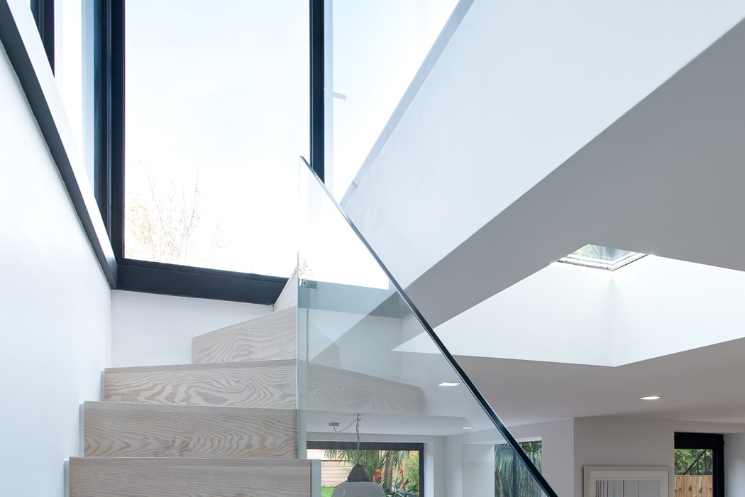 Wooden staircase with glass railing ascending toward large windows in a modern, white interior space with natural light.