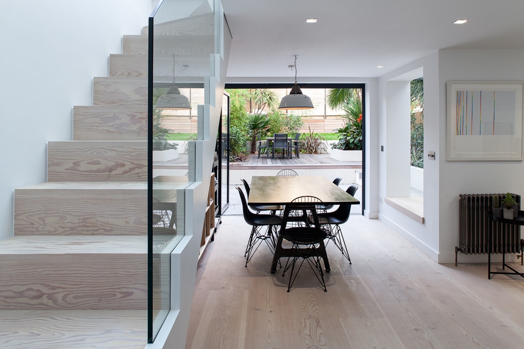 A modern dining area with a wooden table and black chairs sits next to glass-panel stairs, with large sliding doors opening to a patio and garden.