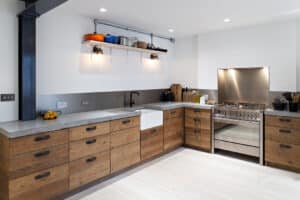 Modern kitchen with wooden cabinets, concrete countertops, a farmhouse sink, stainless steel stove, open shelving with various kitchenware, and minimalist lighting.