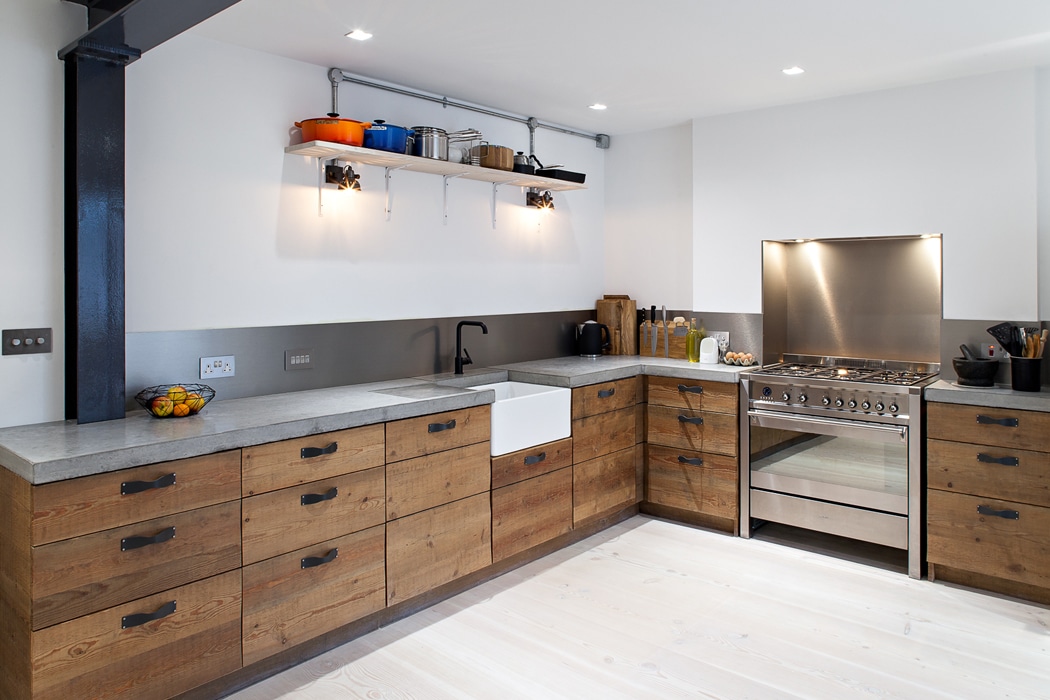 Modern kitchen with wooden cabinets, concrete countertops, a farmhouse sink, stainless steel stove, open shelving with various kitchenware, and minimalist lighting.