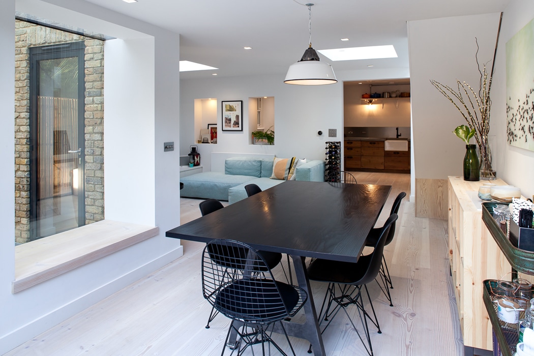Modern dining area with a black table and six chairs, adjacent to a living space with a light blue sofa and open kitchen in a bright room with large windows and white walls.
