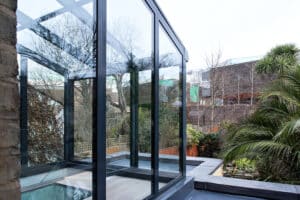A modern glass extension with black frames is attached to a stone building, reflecting trees, plants, and surrounding houses in the garden area.