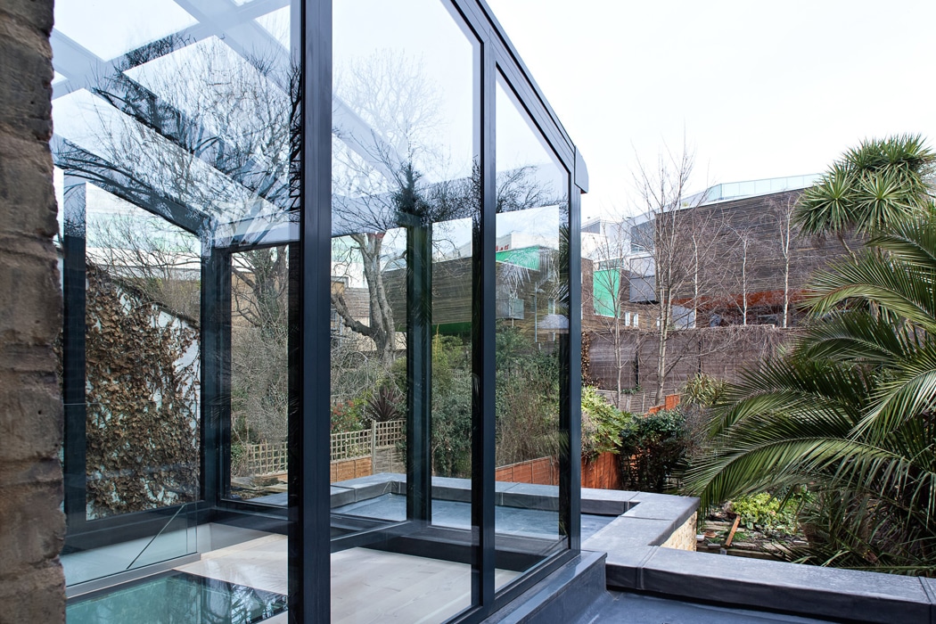 A modern glass extension with black frames is attached to a stone building, reflecting trees, plants, and surrounding houses in the garden area.