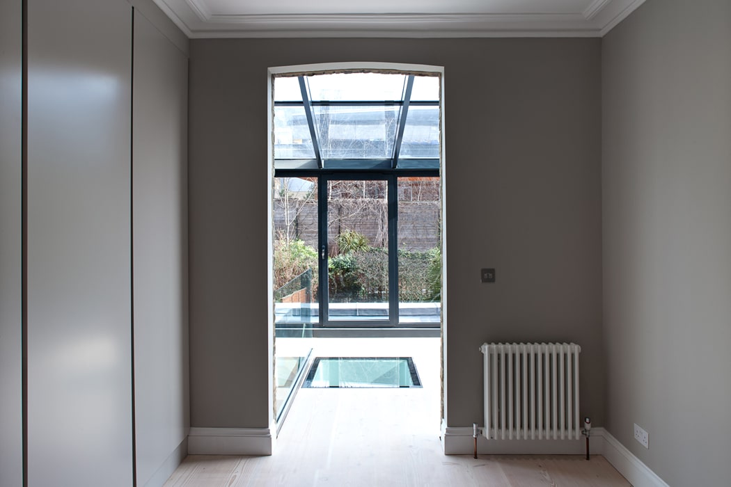A minimalist room with light wood floors, grey walls, a radiator, and a large glass door opening to a patio with a glass roof and views of garden plants outside.