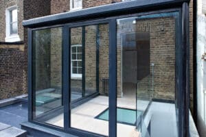 Glass-walled room with sliding doors, flat roof, and skylights, attached to a brick building with white-trimmed windows.