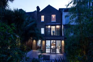 A modern, multi-story brick house with large glass windows and entrances is lit from within at dusk, surrounded by lush greenery.