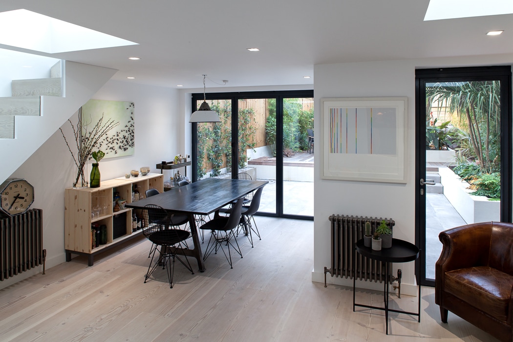 Modern dining area with a black table and chairs, wooden flooring, artwork on white walls, large glass doors leading to a patio, natural light from skylights, and a brown leather chair.