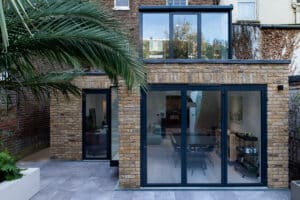 A modern brick townhouse extension with large glass doors and windows opening onto a tiled patio, featuring a palm tree in the foreground.