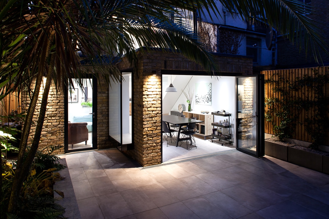 Modern home with brick exterior, large glass doors open to a patio, revealing a brightly lit dining area and interior furnishings at dusk.