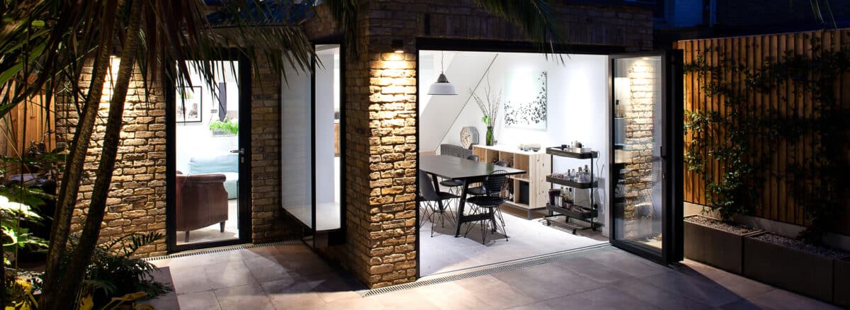 Modern dining area with glass doors, exposed brick walls, and outdoor patio, featuring contemporary furniture and soft indoor lighting.