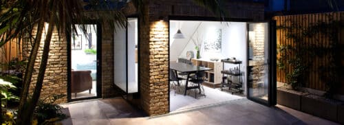 Modern dining area with glass doors, exposed brick walls, and outdoor patio, featuring contemporary furniture and soft indoor lighting.