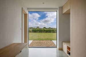 A modern interior hallway with light wood furniture and a large window overlooking a grassy yard, hedge, and blue sky with clouds.