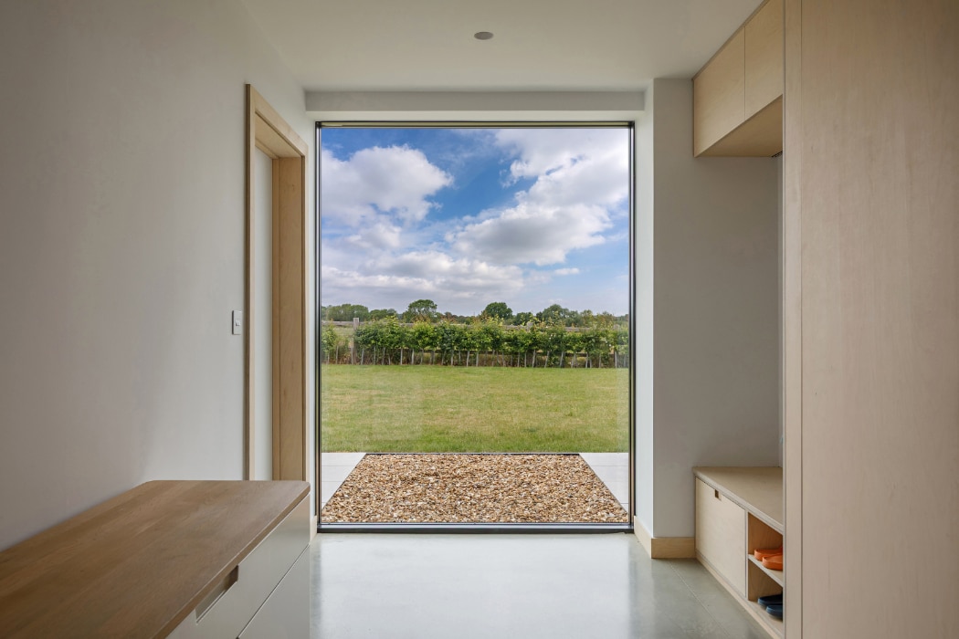 A modern interior hallway with light wood furniture and a large window overlooking a grassy yard, hedge, and blue sky with clouds.