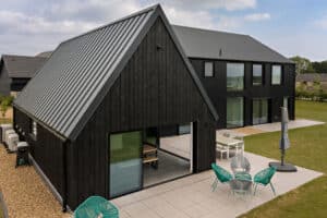 Modern black house with large windows, metal roof, and outdoor patio area featuring turquoise chairs, table, and umbrella on a grassy lawn.