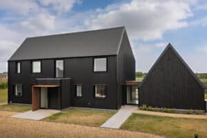 Modern black house with pitched roofs, large windows, and a gravel driveway, set against a cloudy sky and surrounded by grass and open fields.