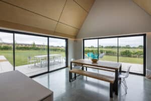 Modern dining area with a wooden table, clear chairs, and large glass doors overlooking a green garden and patio with outdoor seating.