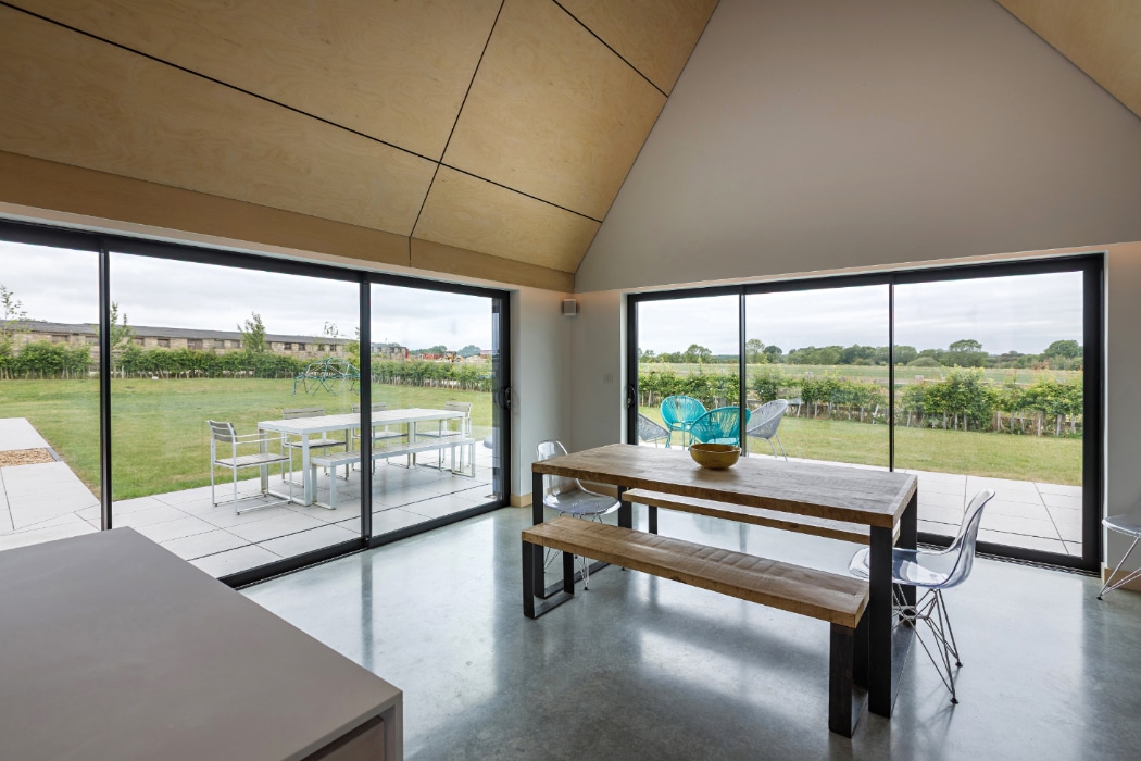 Modern dining area with a wooden table, clear chairs, and large glass doors overlooking a green garden and patio with outdoor seating.