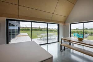 Modern dining area with wooden table and benches, large glass sliding doors, and a view of a patio with outdoor furniture and a green lawn.