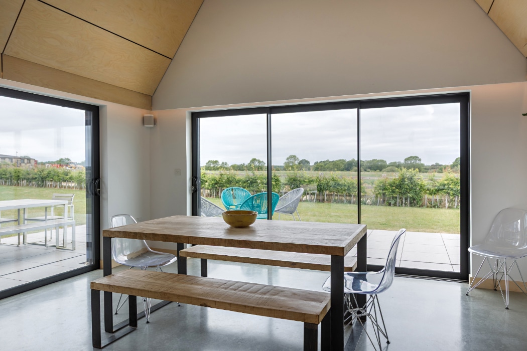 A modern dining area with a wooden table, benches, and clear chairs sits by large sliding glass doors, overlooking a patio and green landscape.