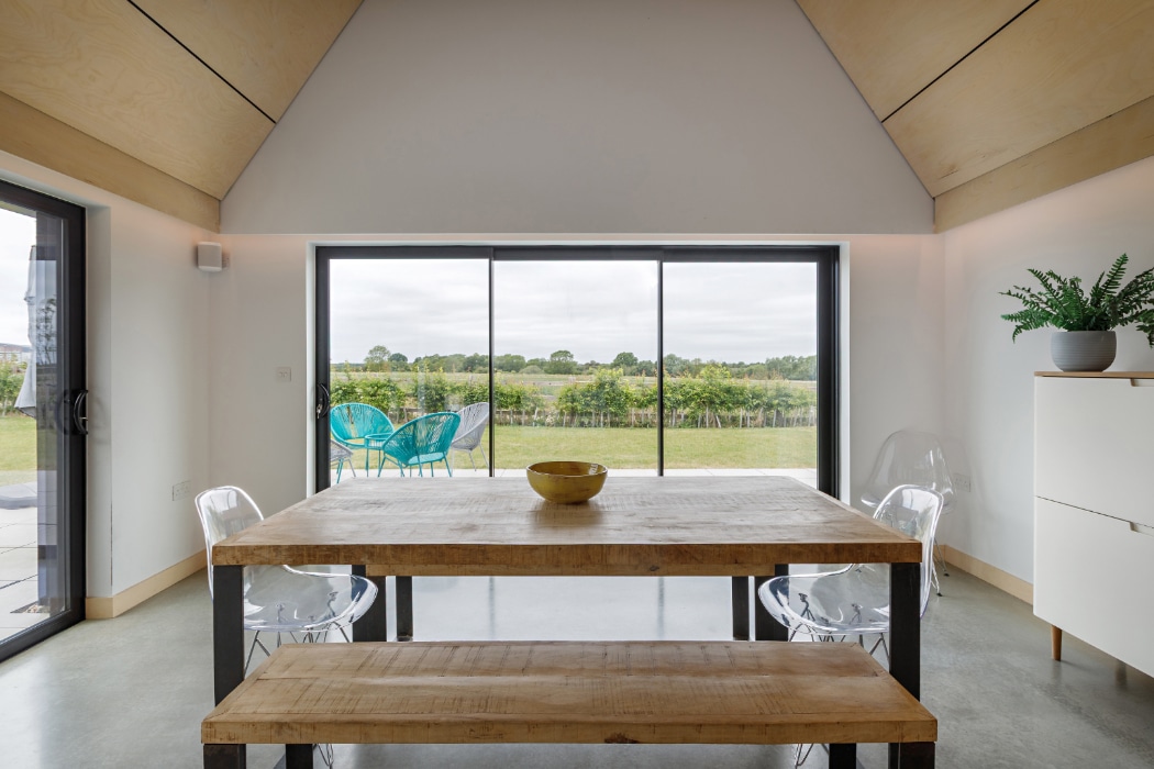 A modern dining area with a wooden table, four transparent chairs, a yellow bowl, and a view of a garden and blue outdoor chairs through large glass doors.