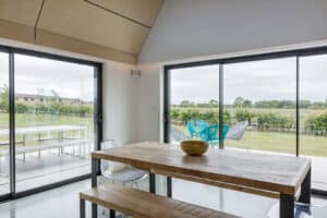 A modern dining area with a wooden table, clear chairs, large glass doors, and views of a grassy outdoor patio and open landscape.