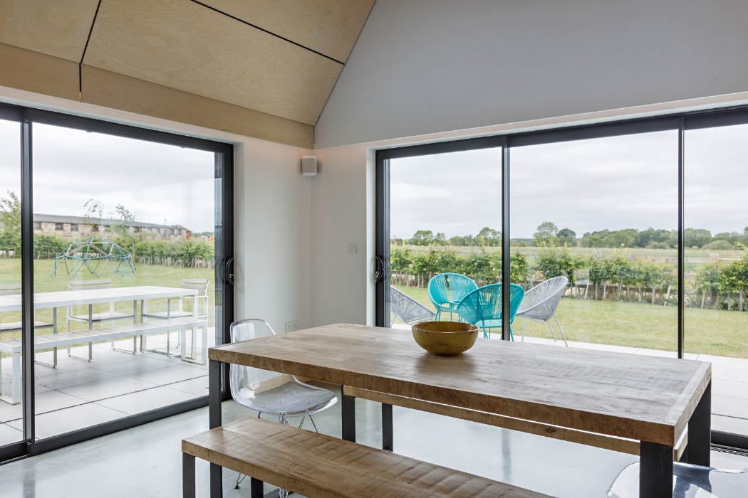 A modern dining area with a wooden table, clear chairs, large glass doors, and views of a grassy outdoor patio and open landscape.