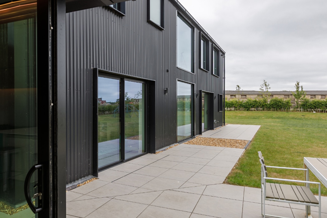 A modern black building with large windows and sliding glass doors opens onto a tiled patio with outdoor seating, overlooking a green lawn.