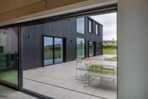 View from inside a modern building looking onto a patio with a white table and benches, and a black, metal-clad exterior wall in the background.