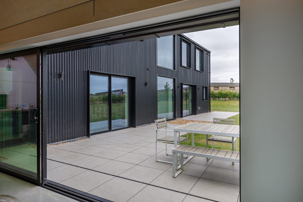 View from inside a modern building looking onto a patio with a white table and benches, and a black, metal-clad exterior wall in the background.