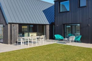 Modern house with dark metal siding and roof, featuring a patio area with white dining furniture and turquoise lounge chairs on a paved surface next to a lawn.